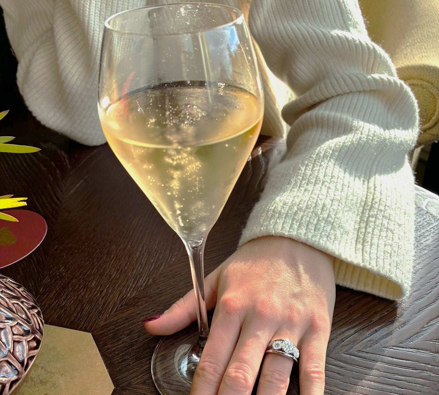 Person holding a glass of white wine with a 3 stone diamond ring on a wooden table
