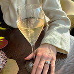 Person holding a glass of white wine with a 3 stone diamond ring on a wooden table