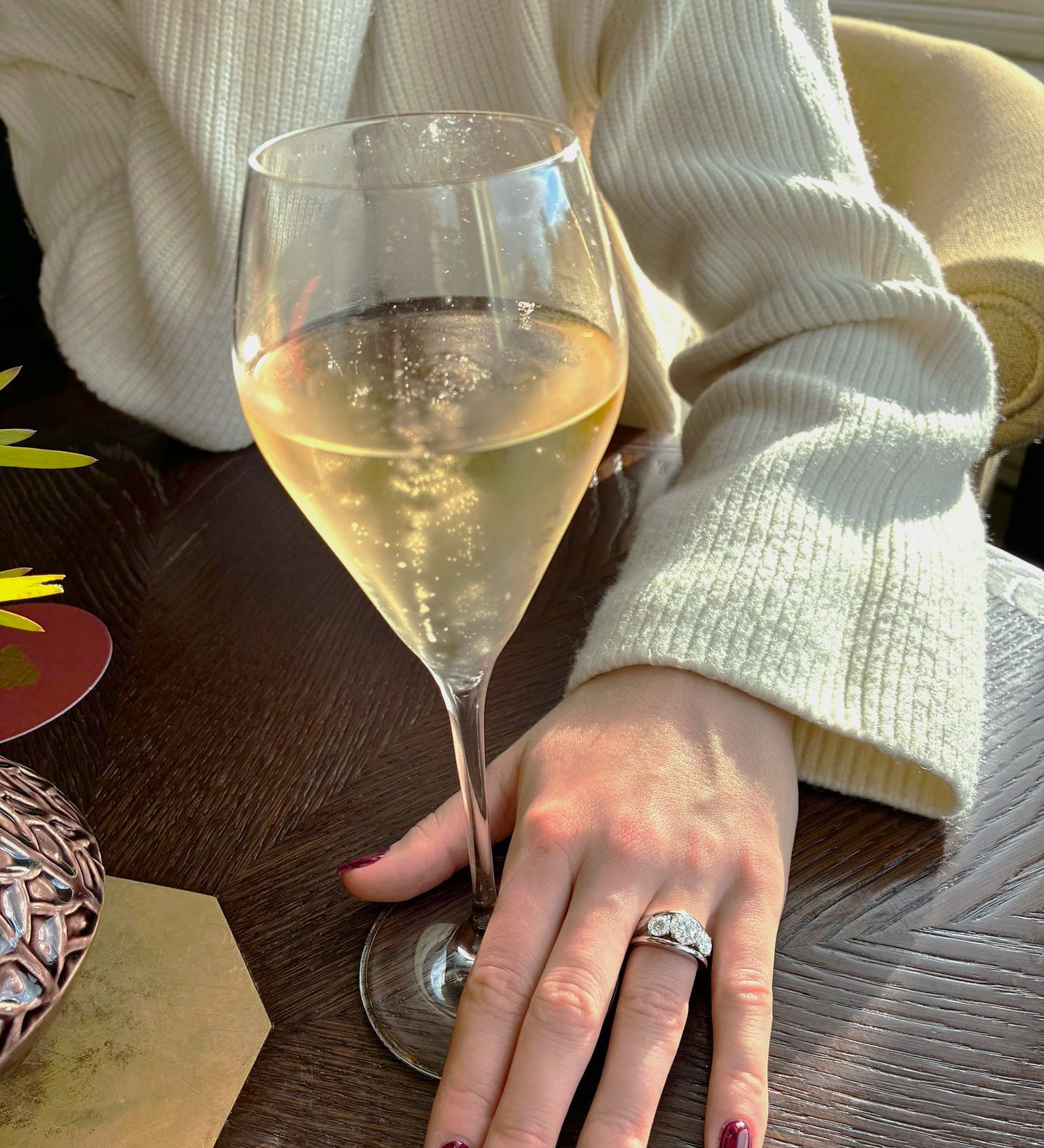 Person holding a glass of white wine with a 3 stone diamond ring on a wooden table