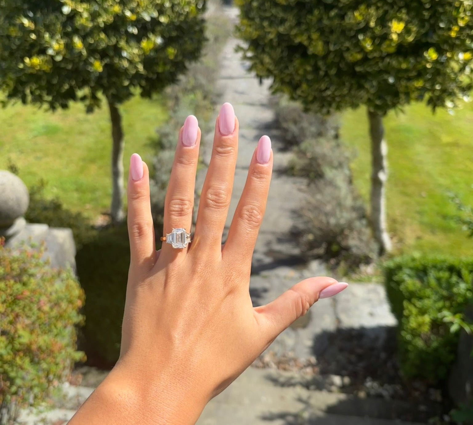 Hand with a 3 stone emerald cut diamond ring on with trees and stone path with greenery